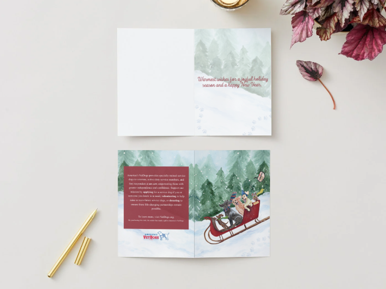 Two cards are displayed on a tabletop: The top card is opened, showing the inside message printed over a pale watercolor background of snow and pine trees. The message reads: “Warmest wishes for a joyful holiday season and a happy New Year." The bottom card is unfolded and shown face-down. The left panel contains a red text box with information about America’s VetDogs, along with the organization’s logo at the bottom. The right panel features the full front illustration of puppies riding in a red sleigh. 