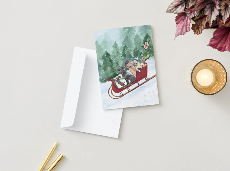 A holiday card rests slightly open on a tabletop beside a plain white envelope. The card’s cover features the playful watercolor illustration of puppies bundled in winter accessories as they ride together in a red sleigh through the snow, set against a backdrop of tall evergreen trees. A lit candle, gold pens, and decorative leaves complete the styled arrangement, giving the image a cozy seasonal feel.
