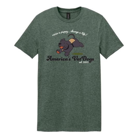 A heather forest green unisex T-shirt featuring a cartoon-style black Labrador puppy joyfully running with a bone in its mouth. The puppy wears a tan and brown training vest. Above the illustration, white text reads “raise a puppy, change a life!” Below, the America’s VetDogs logo is printed in a script font, along with “est. 2003.”